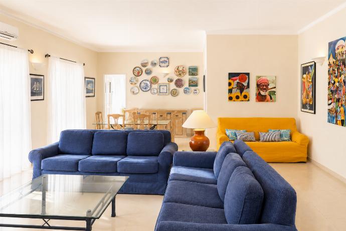 An interior view of a bright, open-plan living and dining room, featuring blue sofas, a yellow accent couch, a glass coffee table, and walls decorated with colorful paintings and decorative plates. . - Villa Green Palms . (Photo Gallery) }}