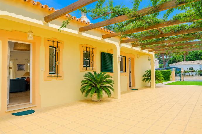 An exterior perspective of a tiled courtyard veranda outside a light yellow villa, featuring an overhead wooden pergola draped with climbing vines and windows fitted with dark green shutters and decorative iron security bars. . - Villa Green Palms . (Photo Gallery) }}