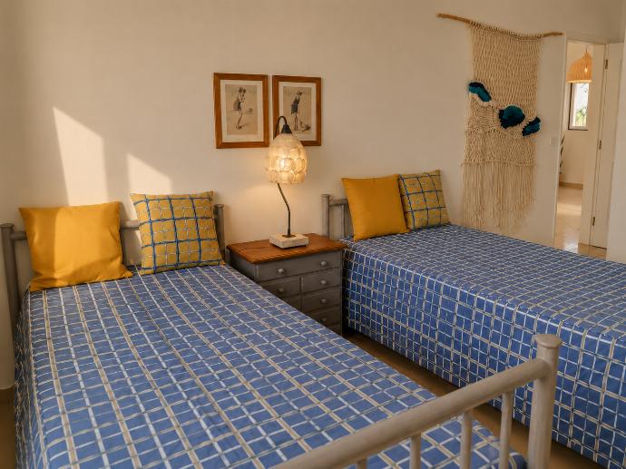 An alternate perspective of the twin bedroom, showcasing the blue-and-yellow plaid bedding, solid yellow pillows, a central grey nightstand with a lamp, and a macrame tapestry near the open doorway. . - Villa Green Palms . (Photo Gallery) }}