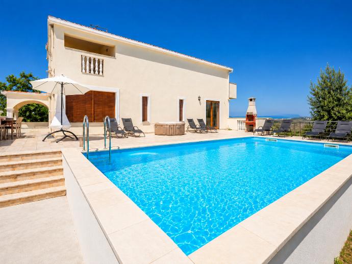 This modern cream-colored villa features a large, crystal-blue swimming pool and a spacious stone patio lined with lounge chairs, a hot tub, and an outdoor barbecue under a clear blue sky. . - Villa Maslina . (Photo Gallery) }}