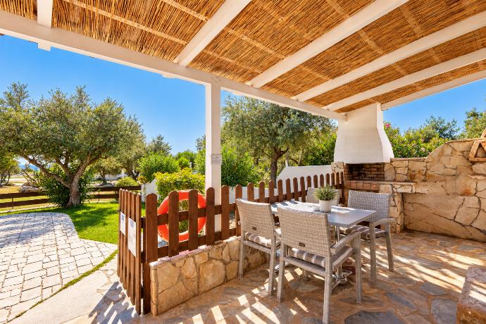 Shaded outdoor dining terrace with rustic wooden furniture, pergola cover, and views toward the private pool and garden.