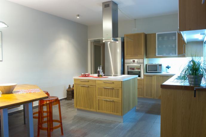 A modern kitchen features wooden cabinetry, a central island with an overhead range hood, and an eat-in dining area with a red cushioned bench and clear chairs. . - Villa Santolina . (Galerie de photos) }}