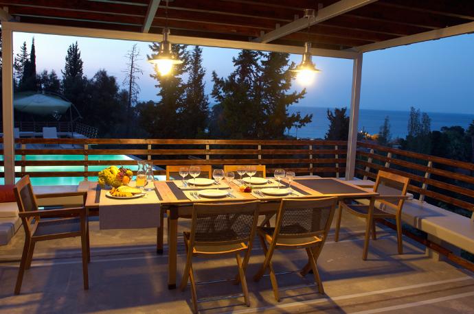 A covered balcony features two clear chairs and a small bistro table under a wooden ceiling, offering a scenic view of green treetops and the distant sea. . - Villa Santolina . (Galerie de photos) }}