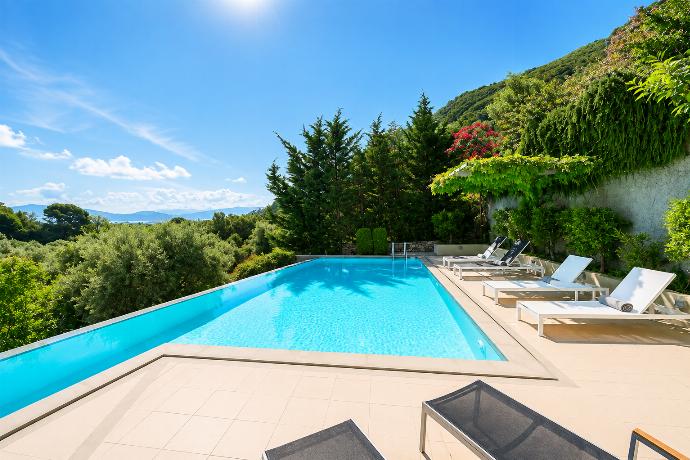 A sun-drenched outdoor swimming pool features lounge chairs arranged on a tiled patio, surrounded by dense green trees and a sweeping view of distant mountains. . - Villa Oleana . (Galería de imágenes) }}