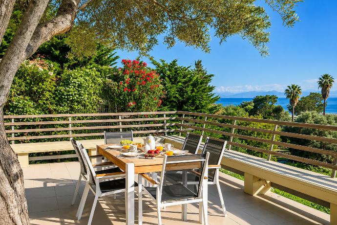 A wooden outdoor dining table is set for breakfast on an open terrace with wooden railings, offering a panoramic view of the sea, palm trees, and lush greenery. . - Villa Oleana . (Galería de imágenes) }}