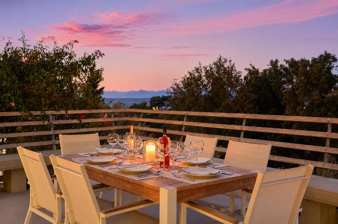 A wooden outdoor dining table is set with wine glasses and a lit candle on a terrace, offering a scenic view of the surrounding trees and sea under a pink sunset sky. . - Villa Oleana . (Galería de imágenes) }}