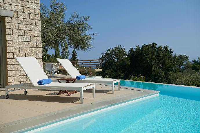 A sun-drenched outdoor swimming pool features lounge chairs arranged on a tiled patio, surrounded by dense green trees and a sweeping view of distant mountains. . - Villa Oleana . (Galerie de photos) }}