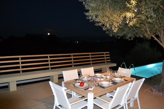 A wooden outdoor dining table is set with wine glasses and a lit candle on a terrace, offering a scenic view of the surrounding trees and sea under a pink sunset sky. . - Villa Oleana . (Galerie de photos) }}