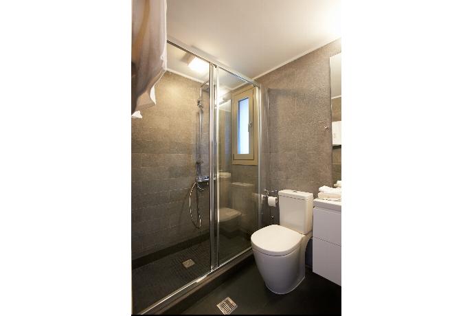 A contemporary bathroom features grey wall tiles, a glass-enclosed walk-in shower, a white vanity with drawers, and a large mirror reflecting an adjacent bedroom. . - Villa Oleana . (Galerie de photos) }}