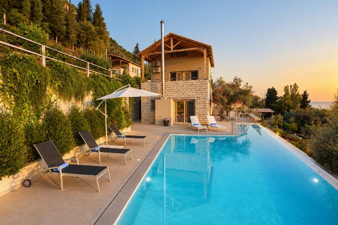 An elevated view showcases a multi-level stone villa nestled into a lush, green hillside, featuring an outdoor swimming pool, patio lounge chairs, and a dining terrace.