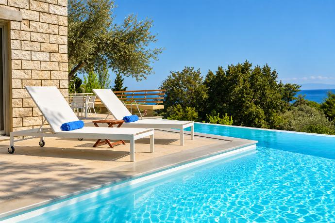 A sun-drenched outdoor swimming pool features lounge chairs arranged on a tiled patio, surrounded by dense green trees and a sweeping view of distant mountains.