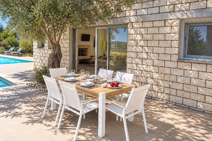 A wooden outdoor dining table sits on a sun-drenched terrace next to a stone villa, featuring a private swimming pool and a lush mountain landscape in the background.