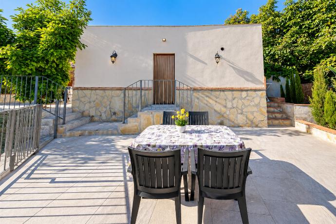 Spacious outdoor dining terrace with comfortable seating, stone detailing, and lush Mediterranean greenery surrounding the villa.