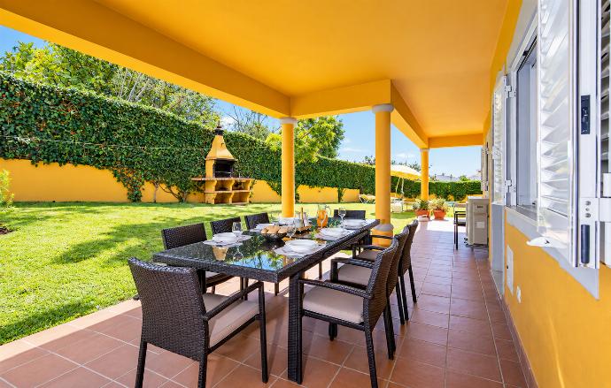 Covered outdoor dining terrace with a large table, comfortable seating, garden views, and shade from the villa veranda.