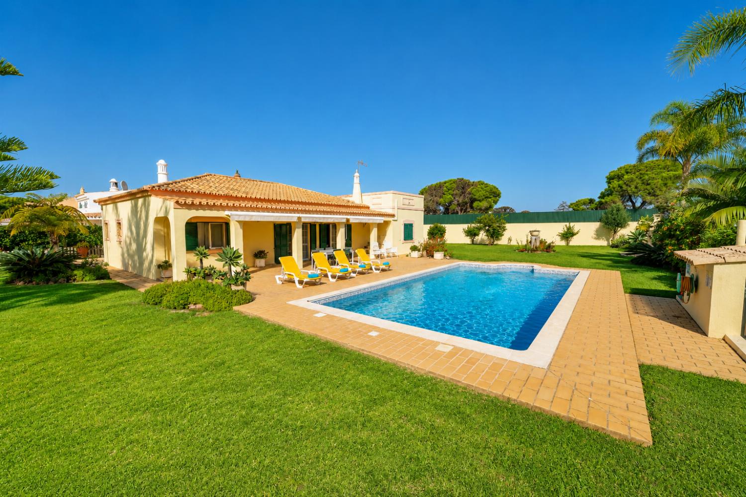 Wide-angle view of a sunny villa backyard featuring a swimming pool, lounge chairs, and a large green lawn.