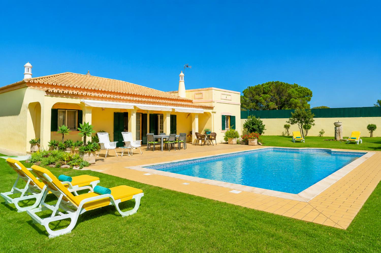 ,A ground-level view of a single-story villa featuring a rectangular swimming pool, a spacious tiled patio with outdoor seating, and a manicured green lawn with yellow loungers under a bright blue sky.