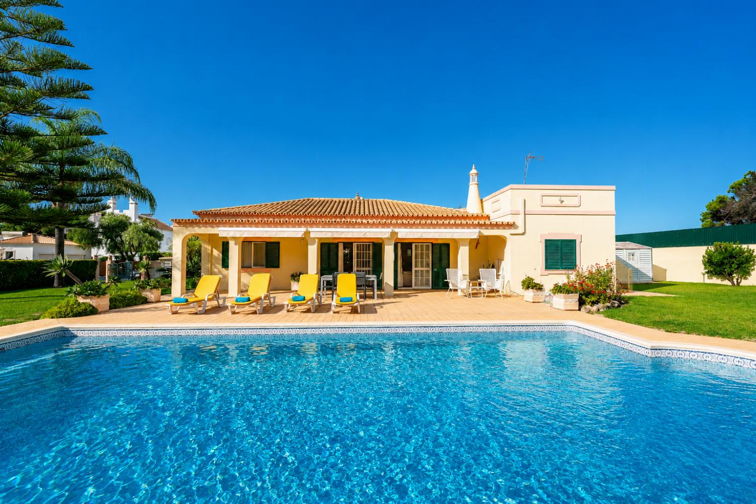 Beautiful single-story villa with a large swimming pool, tiled patio, and yellow lounge chairs.