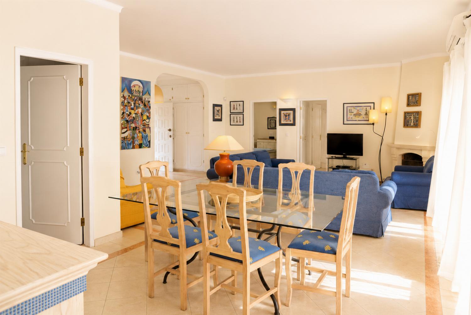 An interior perspective of an open-plan space, centering on a glass-top dining table with light wood chairs, looking through to a living room with blue sofas and a fireplace.