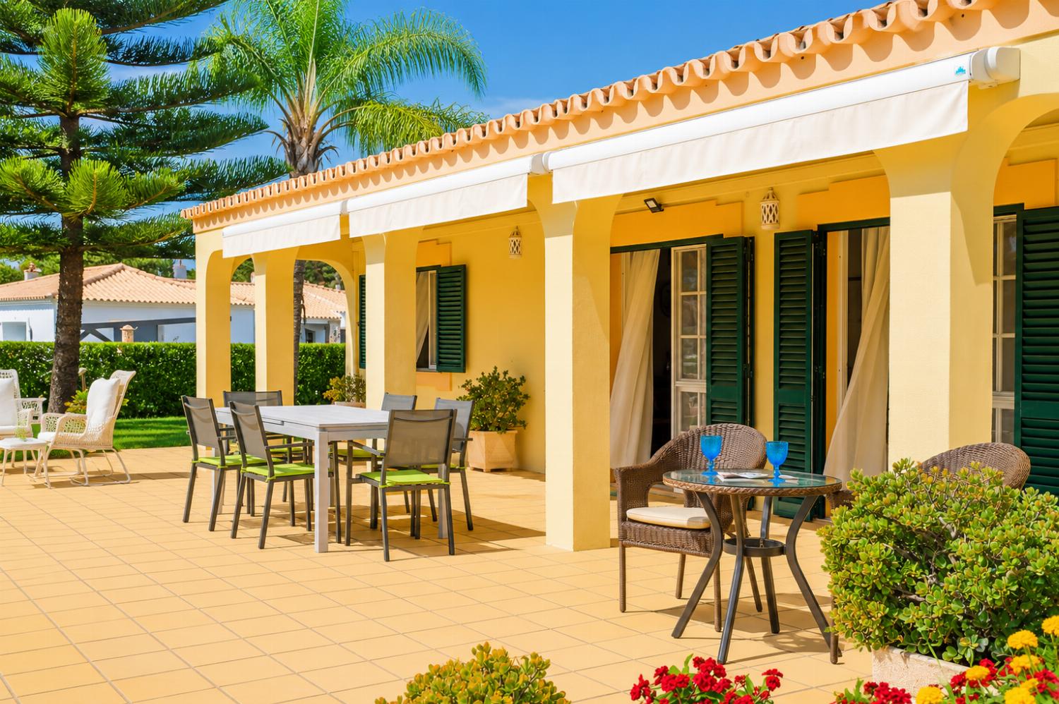 An exterior view of a spacious tiled patio outside a yellow villa, featuring an outdoor dining table with chairs, a smaller wicker bistro set, and retractable white fabric awnings overhead.