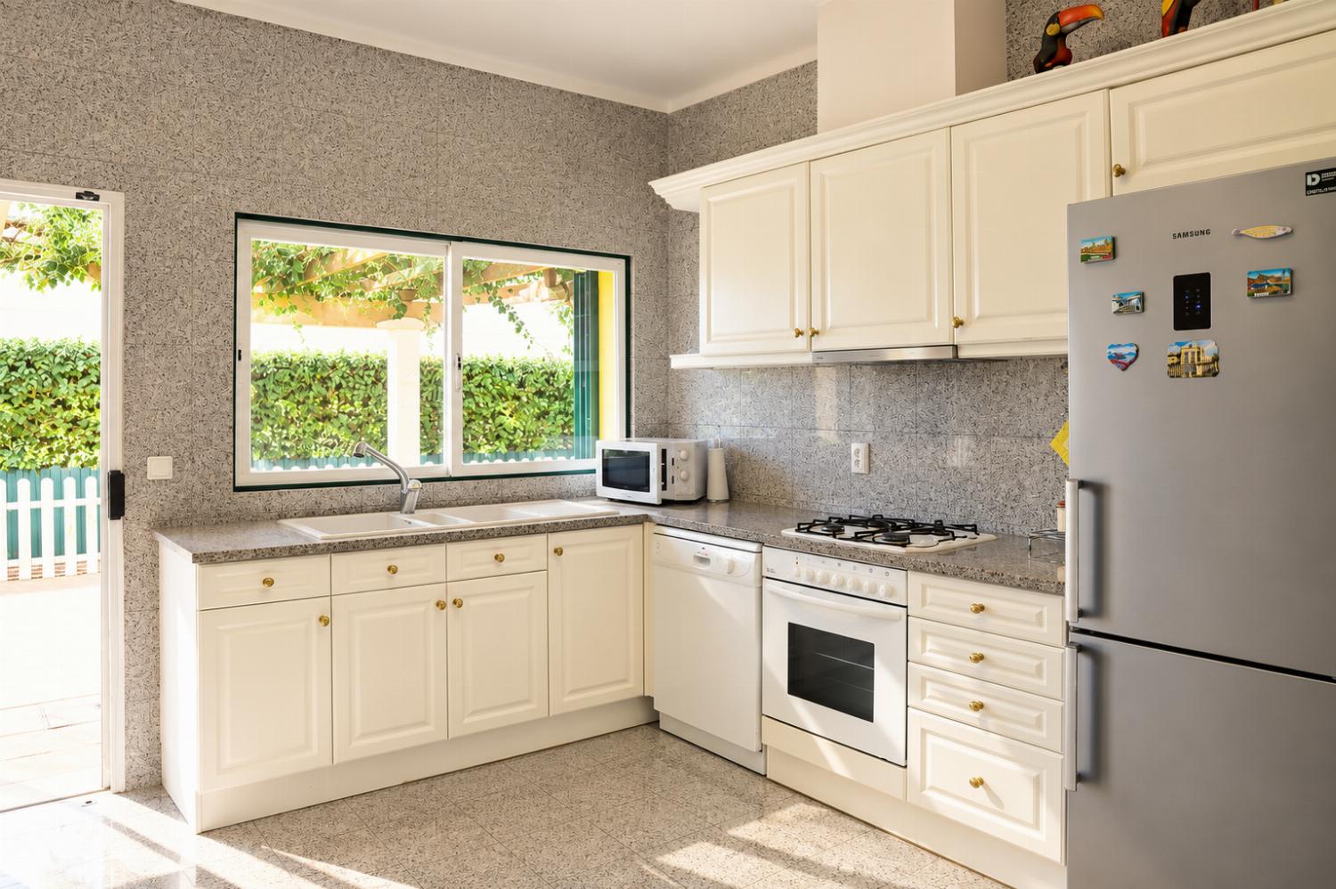 An interior view of a functional kitchen featuring cream-colored cabinets, grey granite-look countertops and wall tiling, integrated white appliances, and a stainless steel refrigerator.