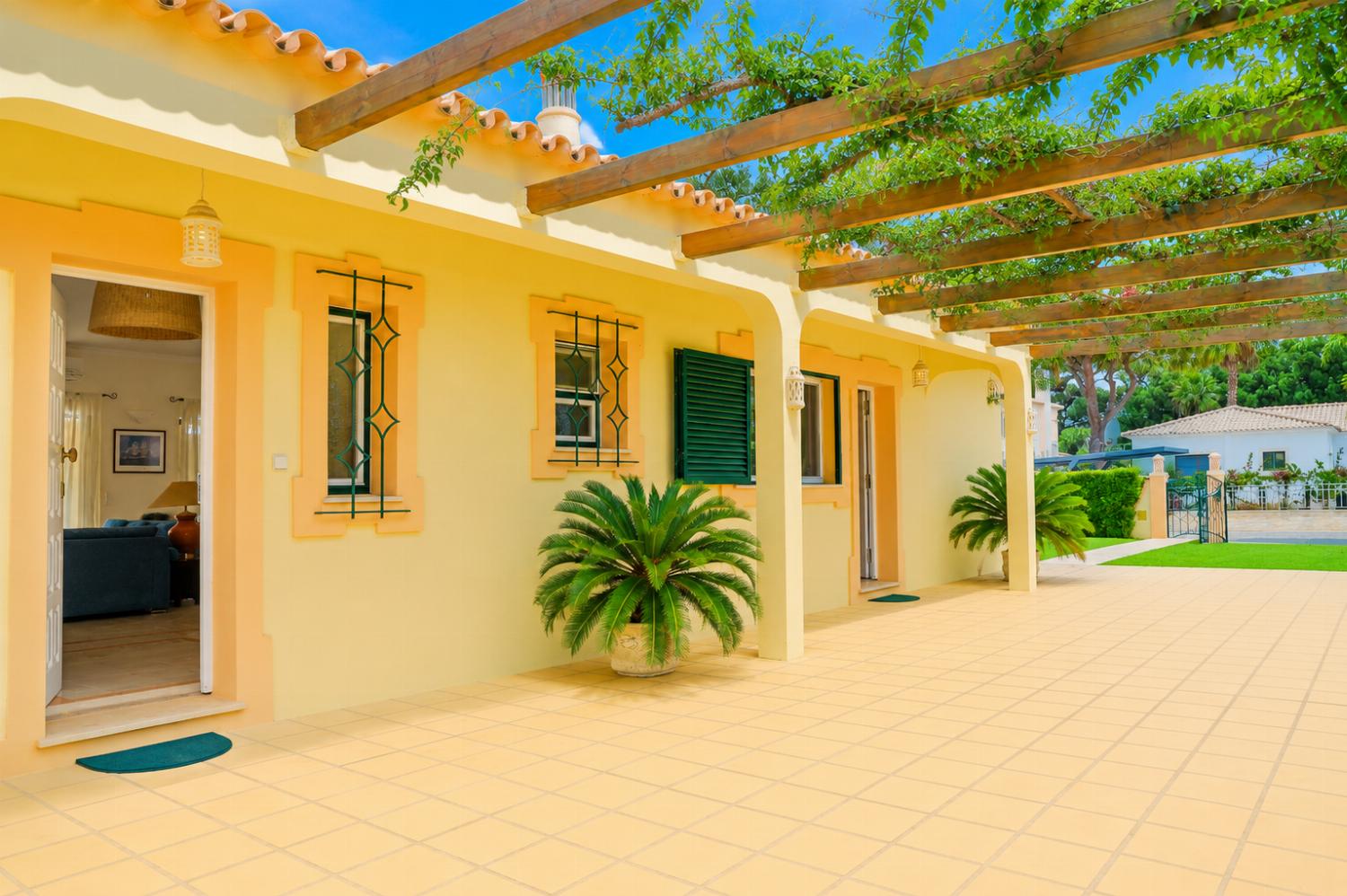 An exterior perspective of a tiled courtyard veranda outside a light yellow villa, featuring an overhead wooden pergola draped with climbing vines and windows fitted with dark green shutters and decorative iron security bars.