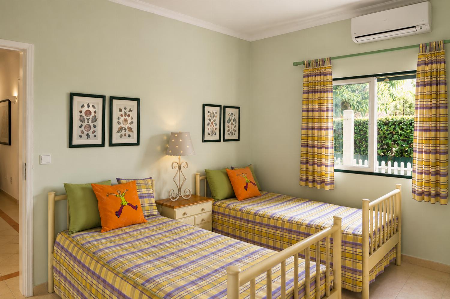 An interior view of a bedroom featuring twin beds dressed in plaid bedding and orange accent pillows, a central nightstand with a lamp, and matching yellow-and-purple curtains framing a window.