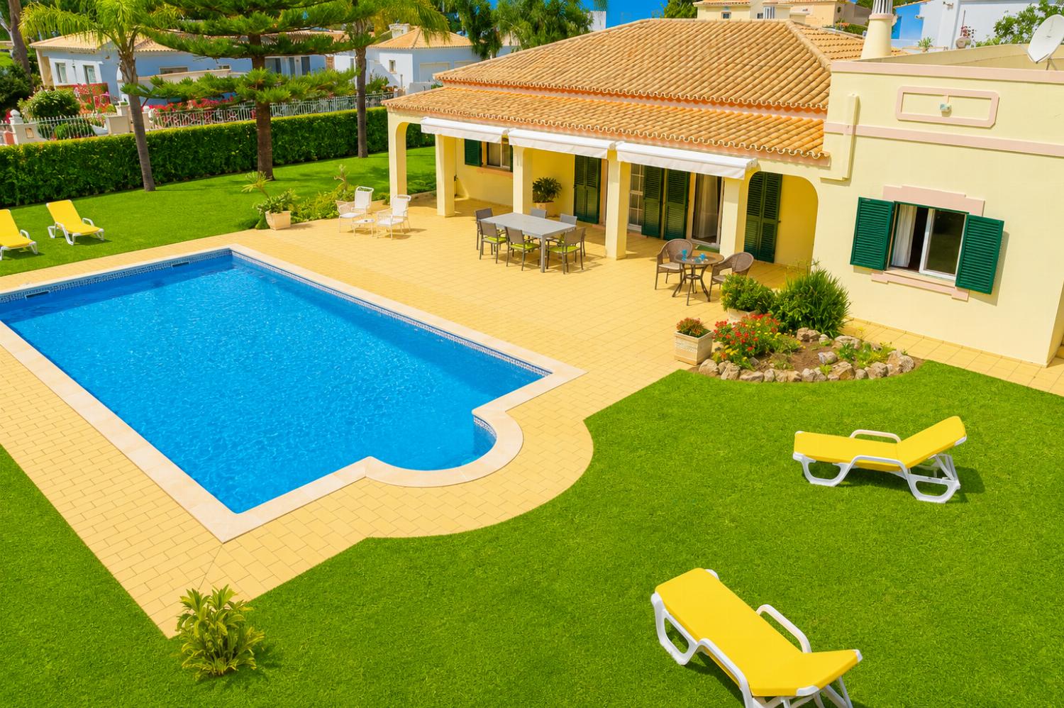 An elevated view of a sun-drenched single-story villa featuring a curved swimming pool, a spacious tiled patio with outdoor dining sets, and a lush green lawn dotted with yellow sun loungers.