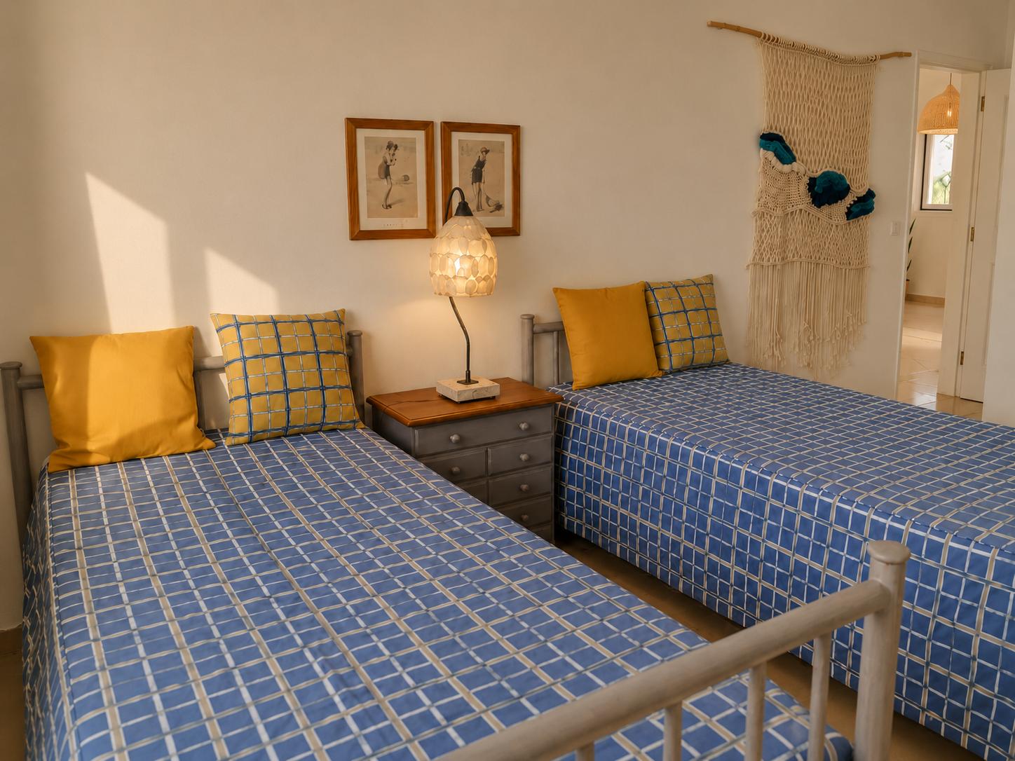 An alternate perspective of the twin bedroom, showcasing the blue-and-yellow plaid bedding, solid yellow pillows, a central grey nightstand with a lamp, and a macrame tapestry near the open doorway.