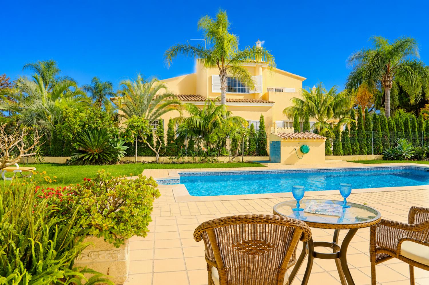 A poolside terrace perspective looking across a swimming pool toward a two-story peach-colored villa, framed by lush tropical palm trees and a wicker seating area in the foreground.