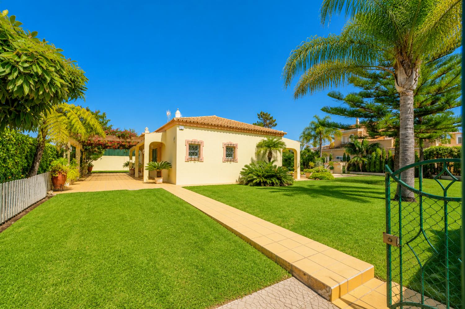 Gated entry view of a single-story villa with a large green lawn, palm trees, and a paved walkway.