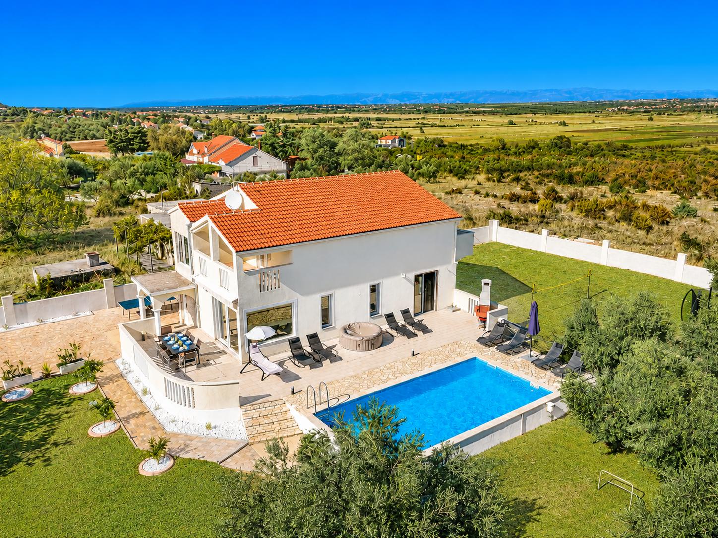 An aerial shot shows a two-story Mediterranean villa with an orange tiled roof, a backyard swimming pool, and an outdoor dining area overlooking a vast rural landscape.