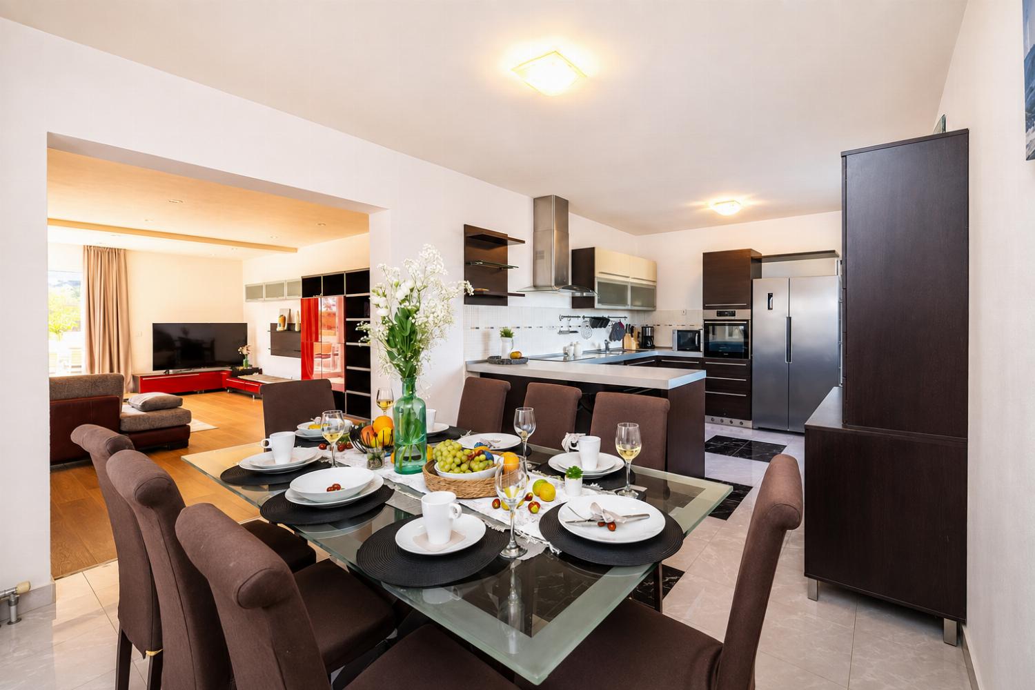 A modern open-plan dining and kitchen area features a glass-top dining table set for six with brown upholstered chairs, opening up to a contemporary kitchen with dark wood cabinets and stainless steel appliances.