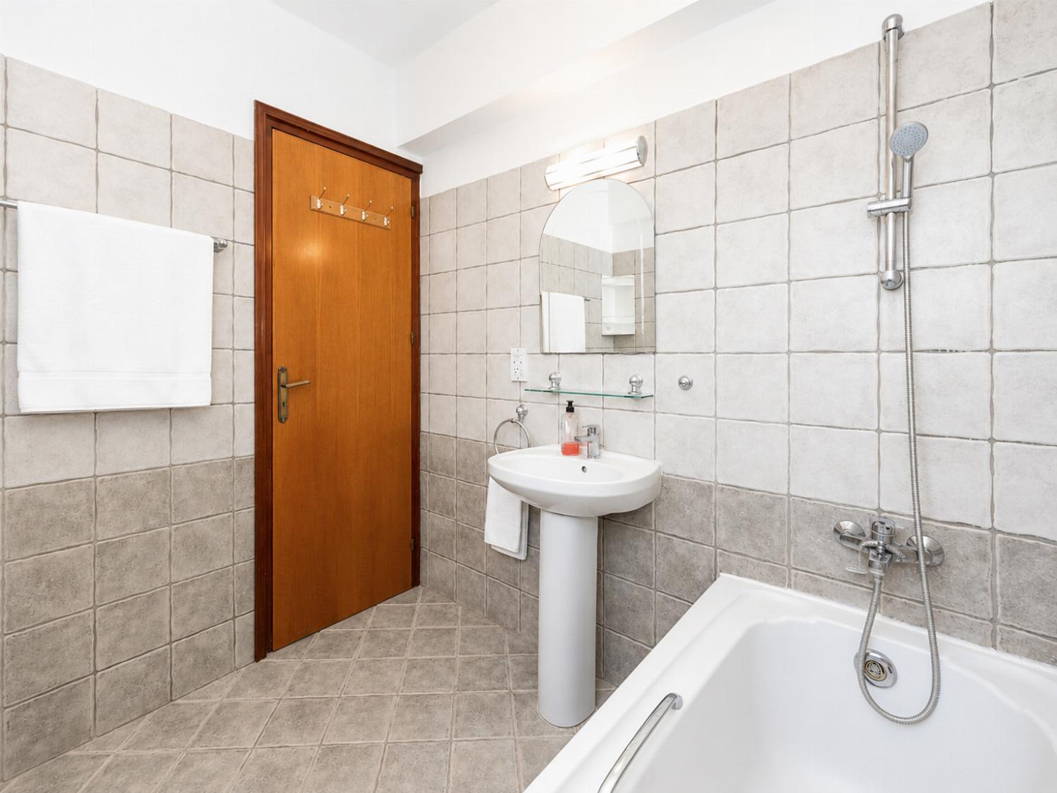 A classic tiled bathroom featuring a corner bathtub, pedestal sink with mirror, toilet, and vibrant yellow accent towels.