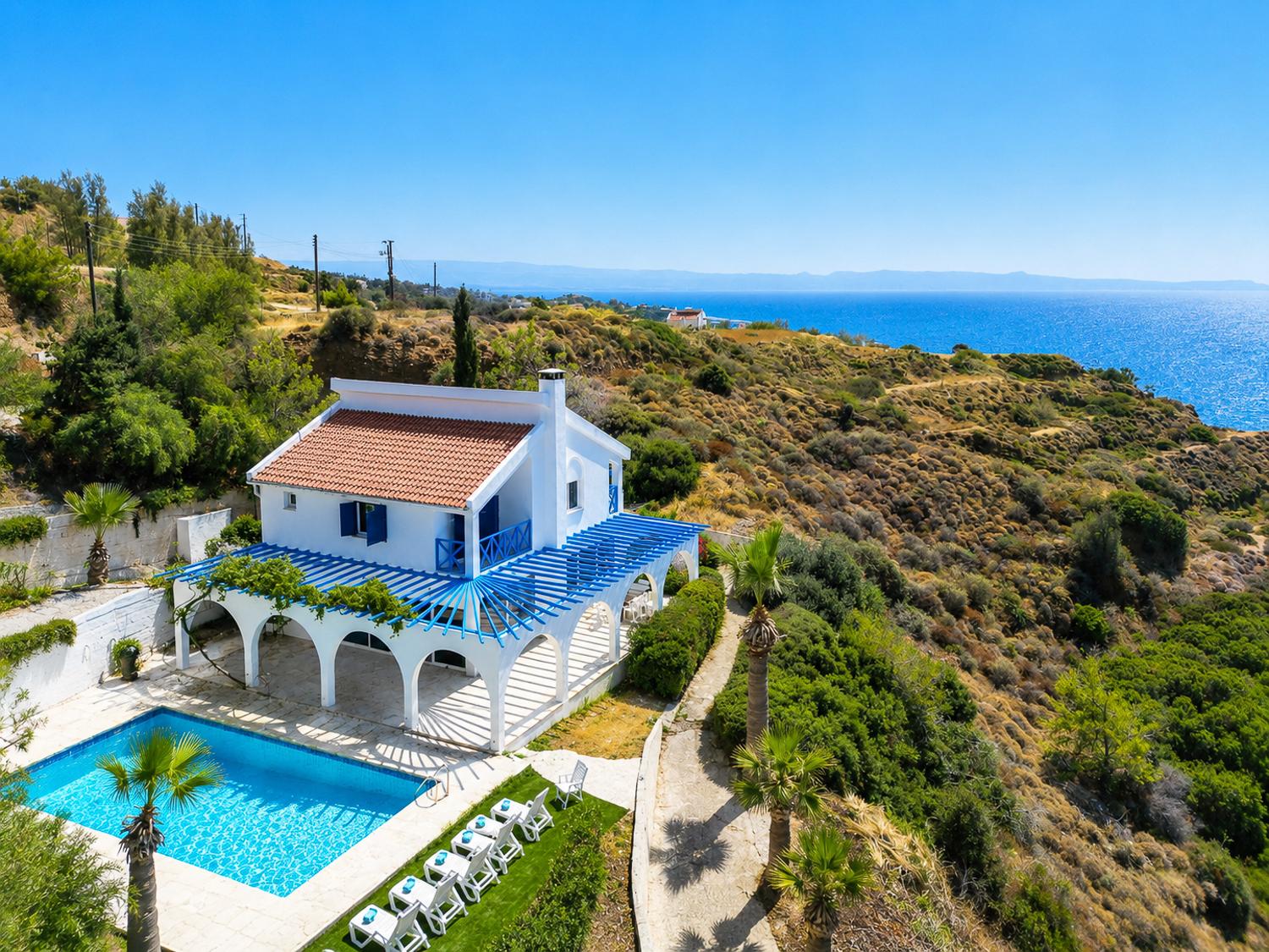 Aerial seafront villa with a private pool, white sun loungers, and stunning coastal views.