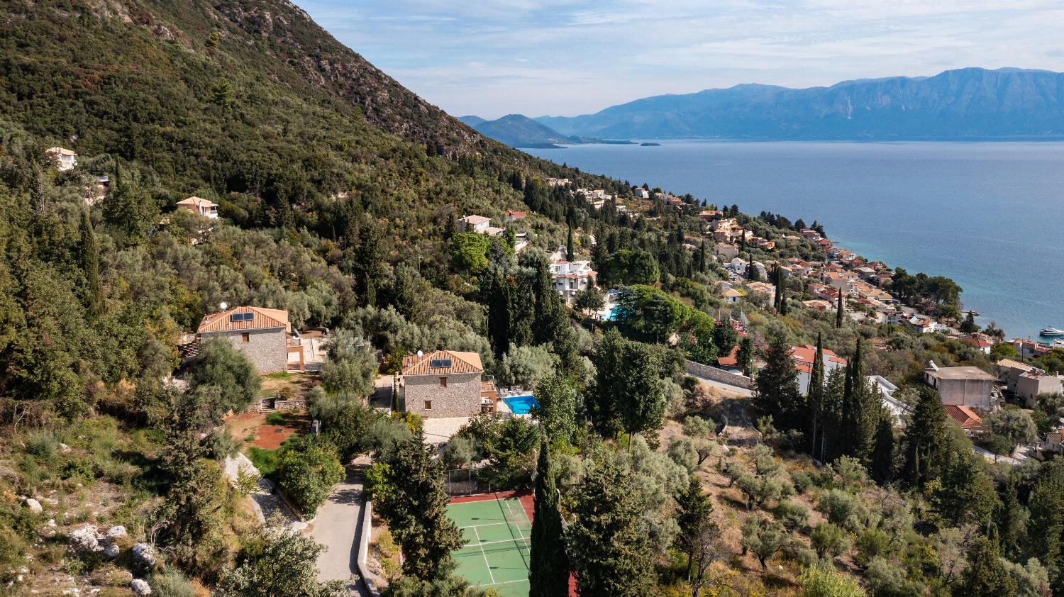 Wide aerial view of hillside stone villas and a tennis court overlooking a coastal village and sea.