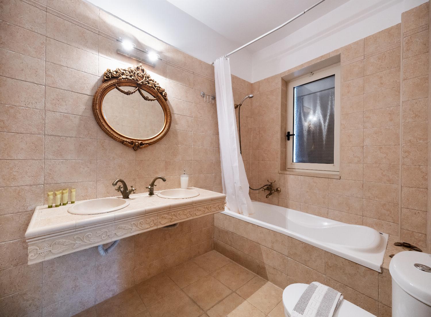 Beige tiled bathroom featuring a built-in bathtub, a vanity sink, and an ornate gold mirror.