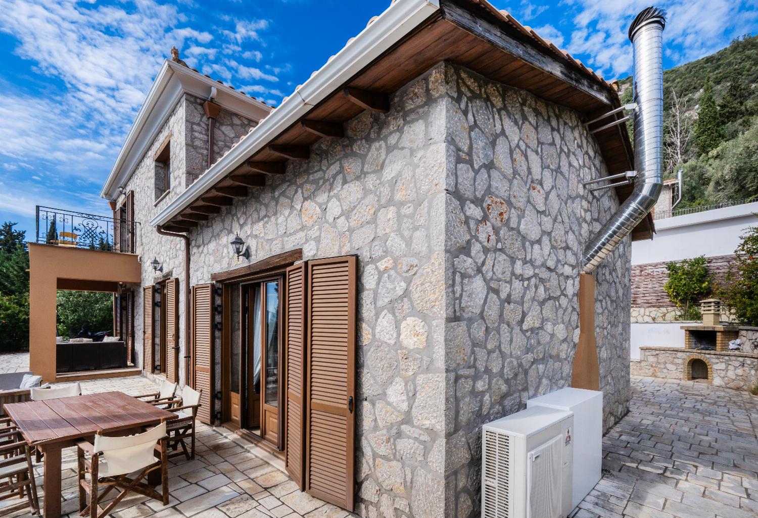Stone villa terrace with a wooden dining set, window shutters, and a stone barbecue.
