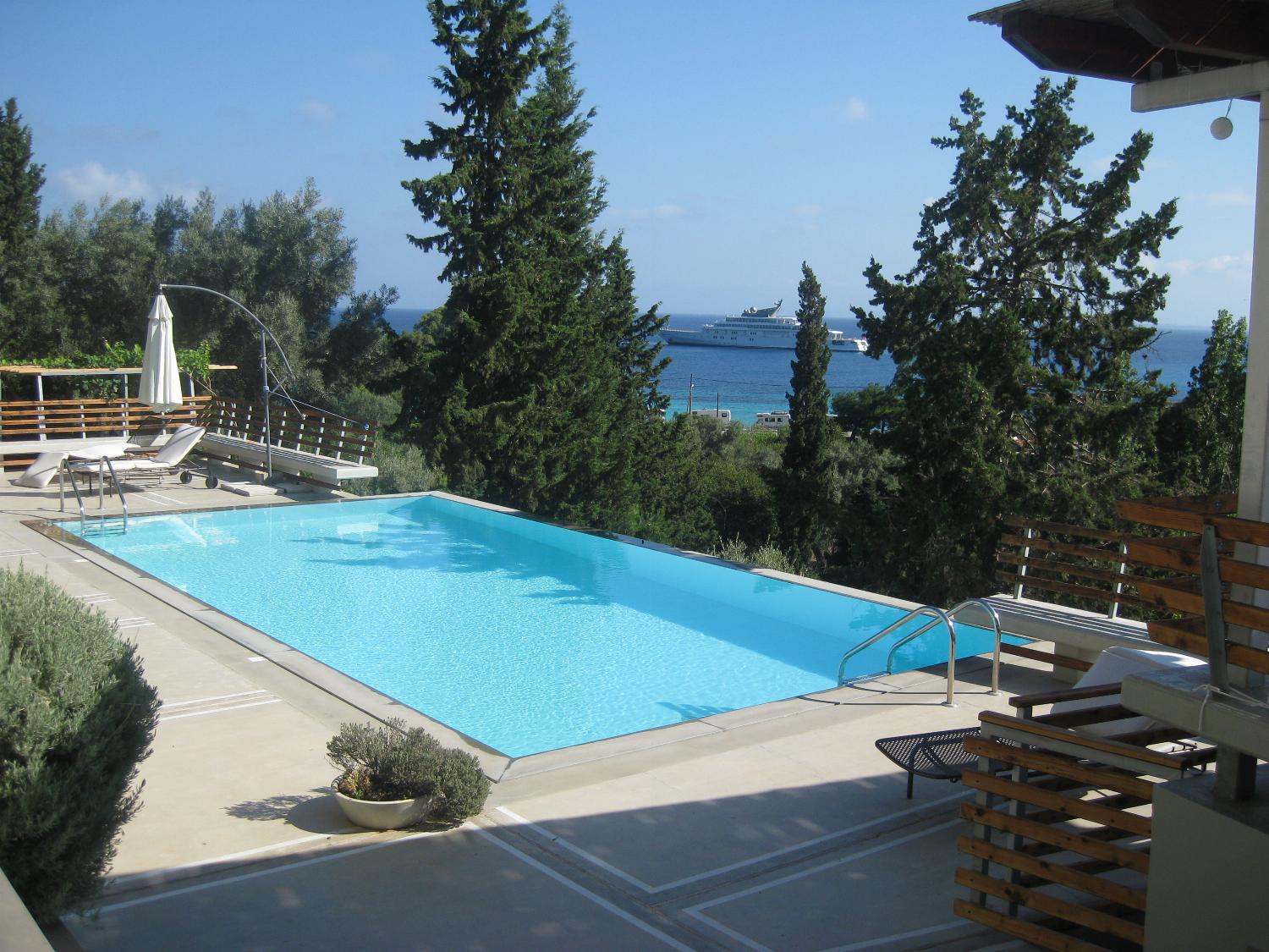 A sunlit outdoor swimming pool features lounge chairs and tall cypress trees overlooking a scenic sea view.