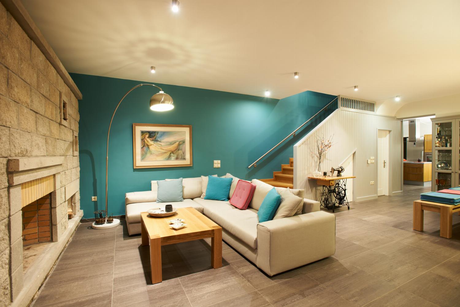 A spacious open-plan living room features a grey sectional sofa against a vibrant turquoise wall, an adjoining wooden dining set, and dark tiled flooring.