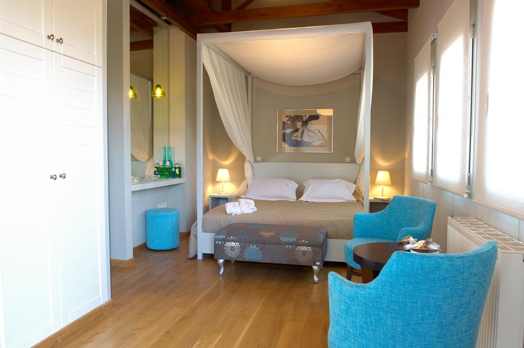 A bright bedroom features a four-poster bed with white draping, exposed wooden ceiling beams, and a pair of vibrant blue accents including a plush armchair and a matching vanity stool.