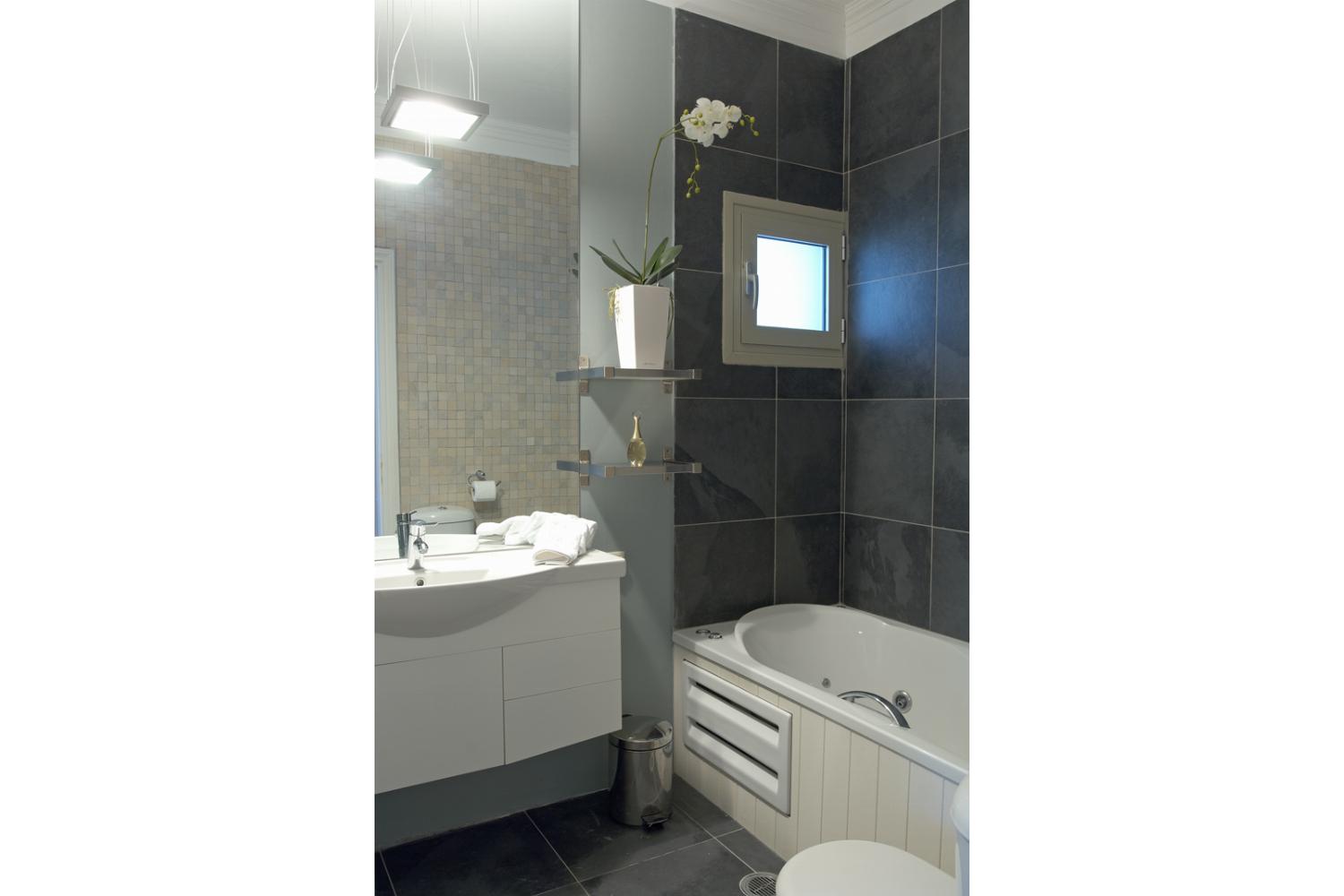 A contemporary bathroom features dark slate-look tiling over a jetted bathtub paired with a white floating vanity and a large wall mirror.