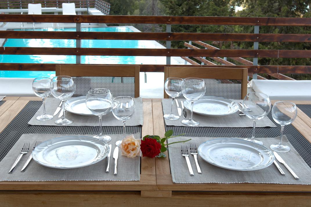 A wooden outdoor dining table is set with lit candles on a covered terrace, offering a scenic view of the coastline during a warm sunset.