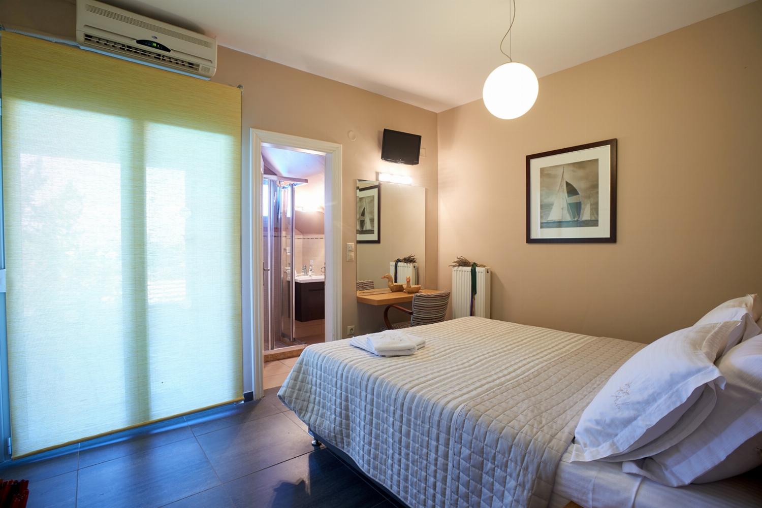 A bright bedroom features a double bed with coral-colored pillows, dark tile flooring, and large glass doors that open directly onto a scenic balcony with a sea view.