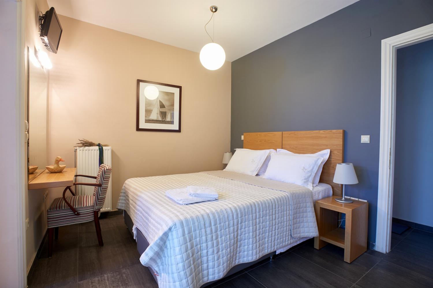 A modern bedroom features a double bed with a wooden headboard, a textured grey accent wall, and open doors leading out to a sunlit balcony with a coastal view.