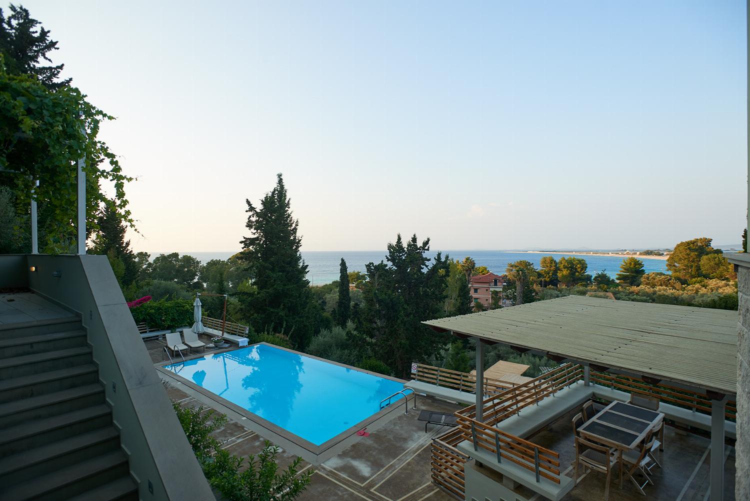 An elevated view highlights the outdoor swimming pool surrounded by a concrete deck, wooden benches, and green trees extending toward the coastline.