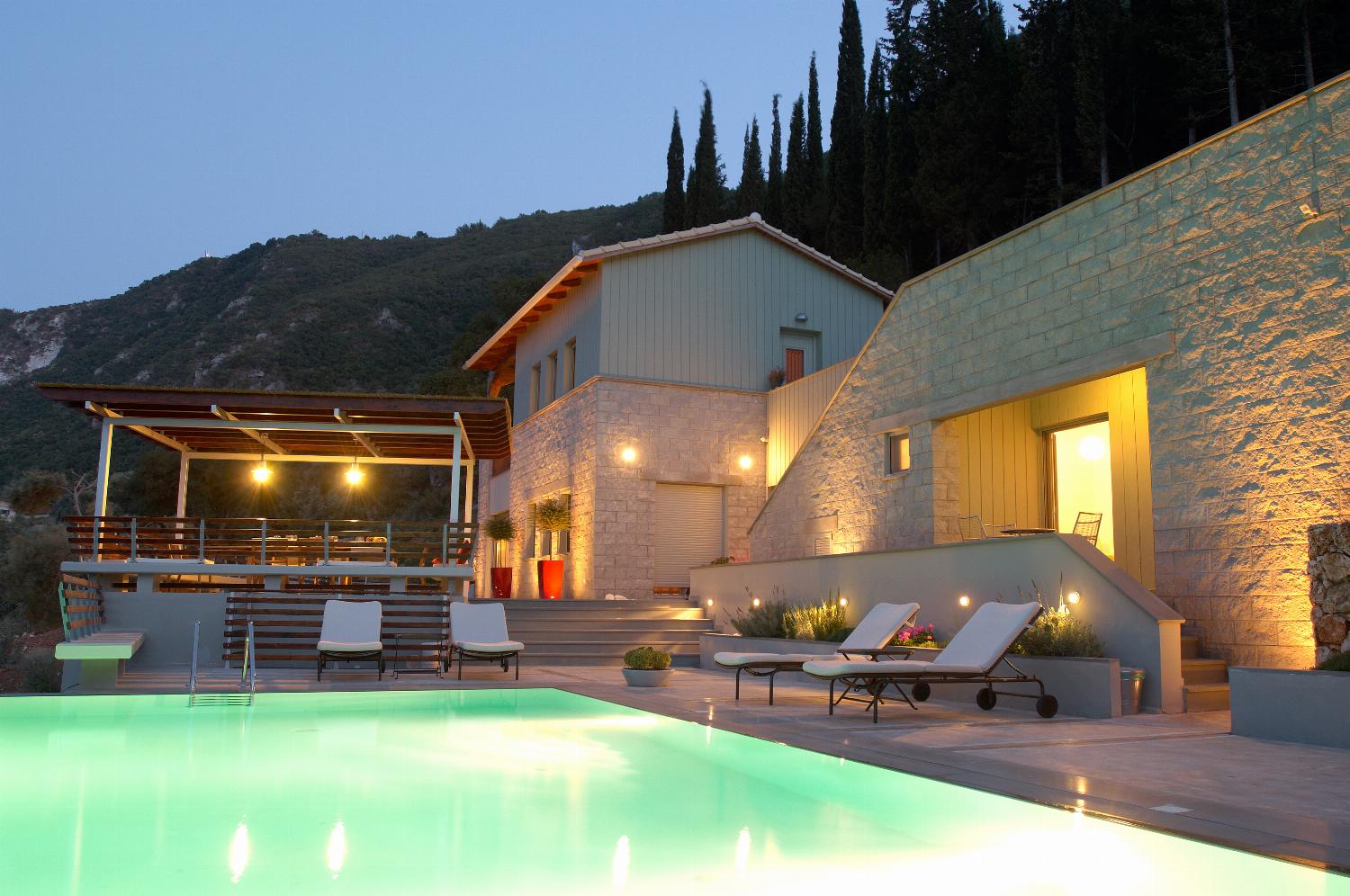 An elevated evening view showcases a multi-level hillside villa illuminated at dusk, featuring a glowing outdoor swimming pool, terrace spaces, and surrounding green forest.