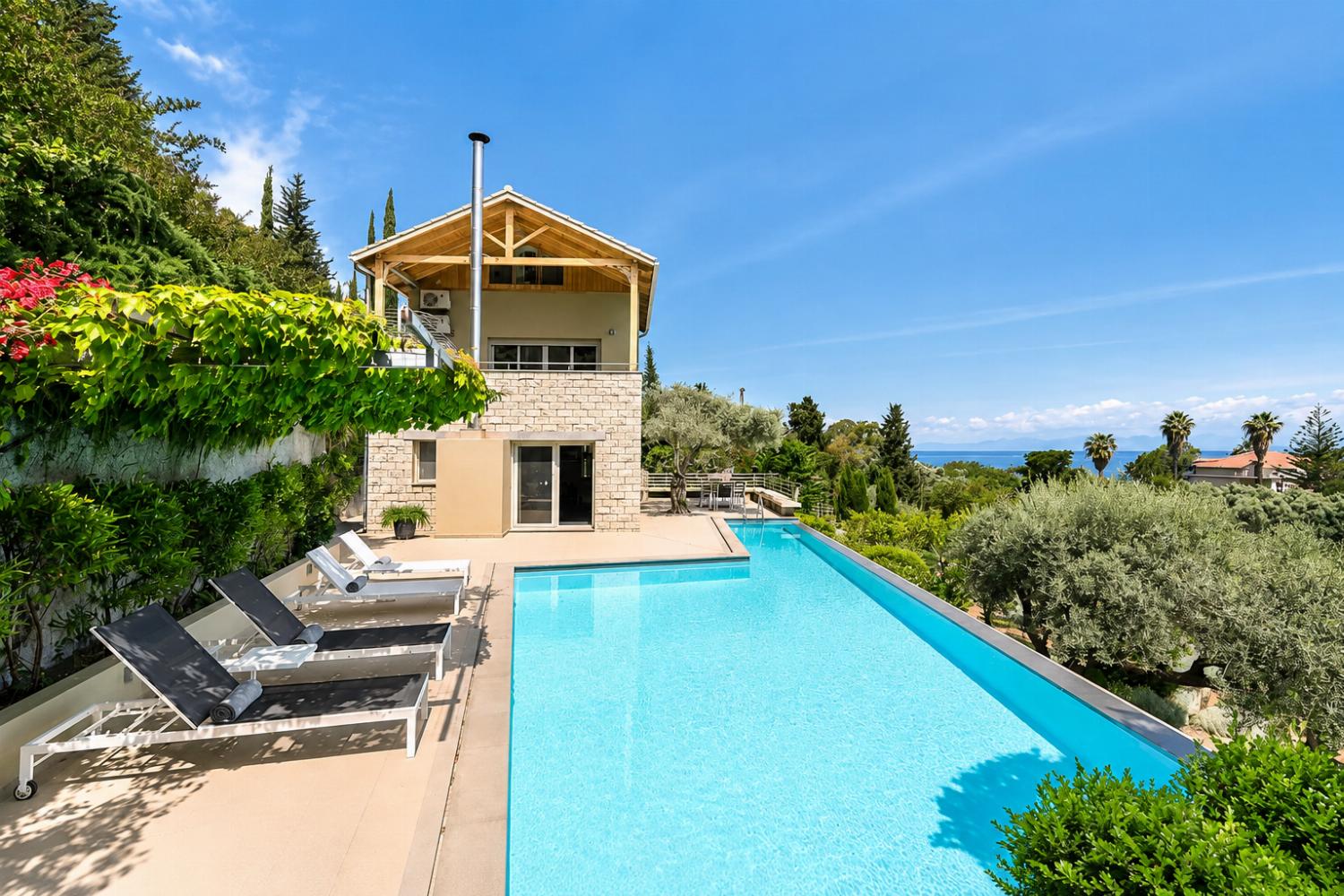 A patio view showcases lounge chairs lining a bright blue swimming pool next to a multi-story stone villa, offering a panoramic outlook toward the distant sea.