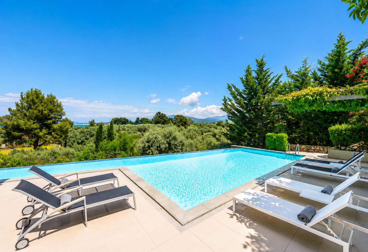 An expansive outdoor swimming pool is surrounded by lounge chairs on a spacious tiled deck, overlooking a vast landscape of green trees under a bright blue sky.
