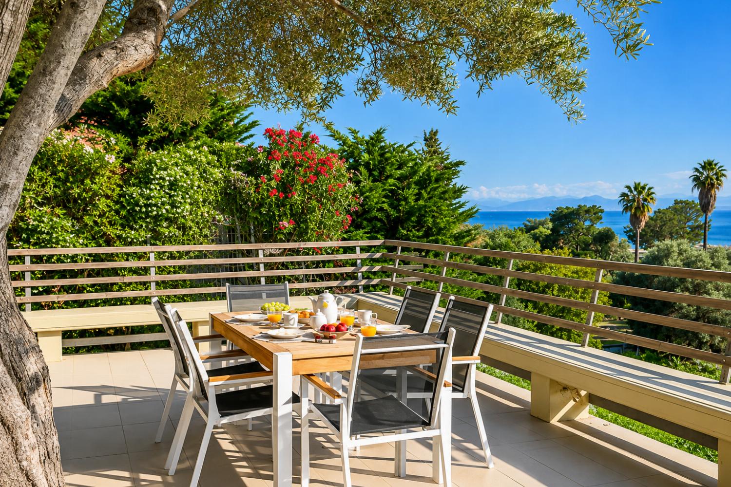 A wooden outdoor dining table is set for breakfast on an open terrace with wooden railings, offering a panoramic view of the sea, palm trees, and lush greenery.