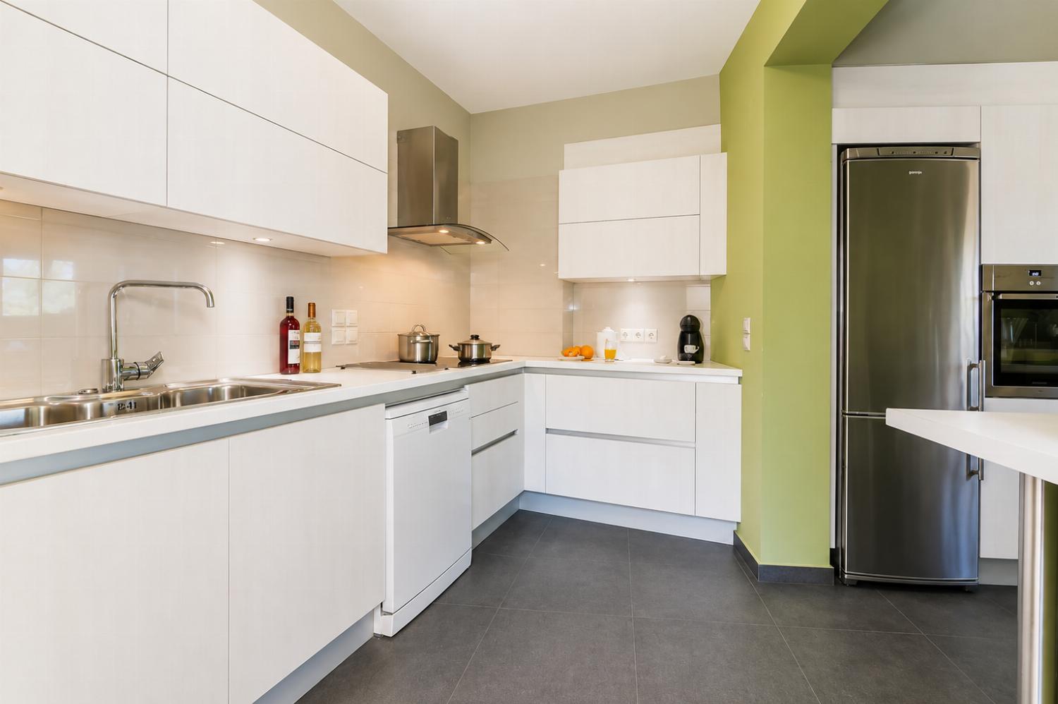 A modern kitchen features sleek white cabinetry, integrated stainless steel appliances, dark floor tiles, and a light green accent wall.